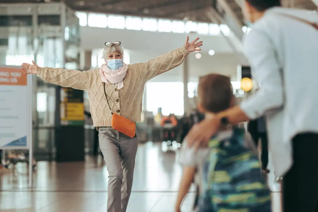Senior woman with open arms waiting to greet family at airport wearing face mask