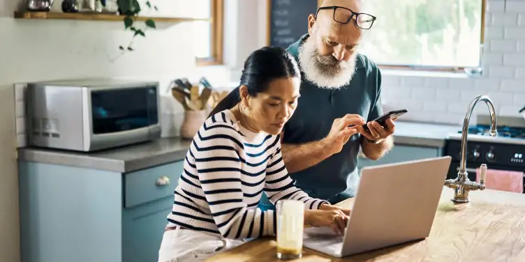 Mature couple sat at a kitchen table looking at a laptop booking a holiday.