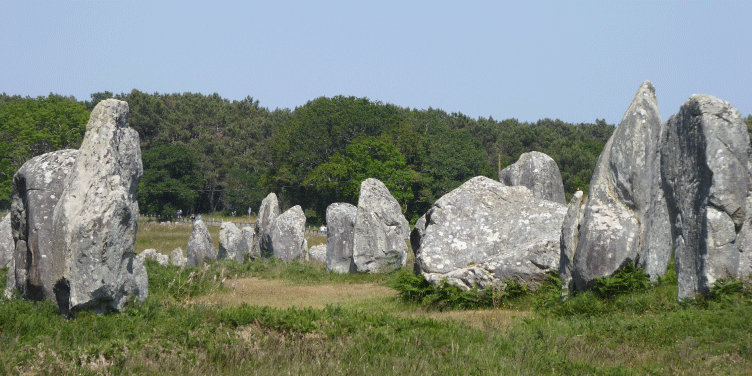 Tourists gather around neolithic megaliths placed in a field on a bright sunny day.