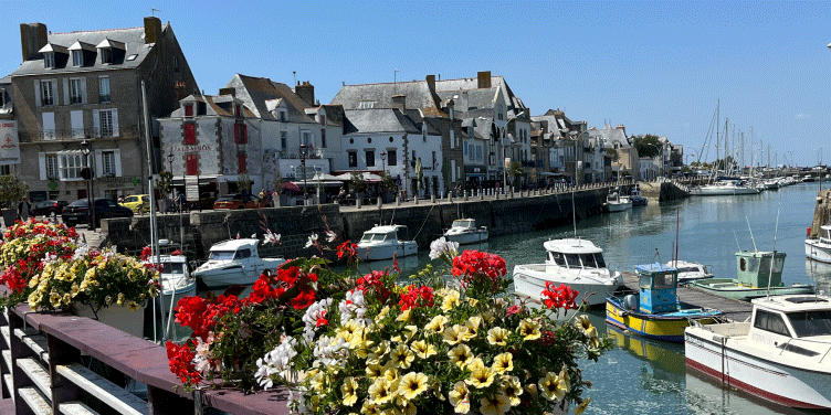 Vibrant flowers perched on the harbour in Le Croisic, Brittany. A few boats have moored up and the sun is shining down on the water.