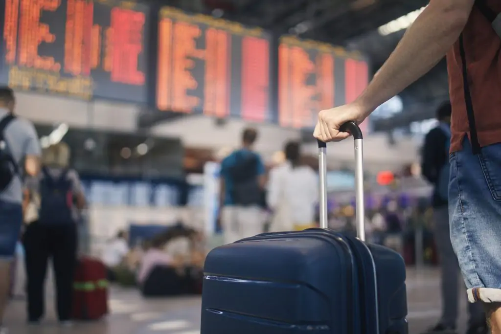 Hand of man while holding suitcase against arrival and departure board at airport.