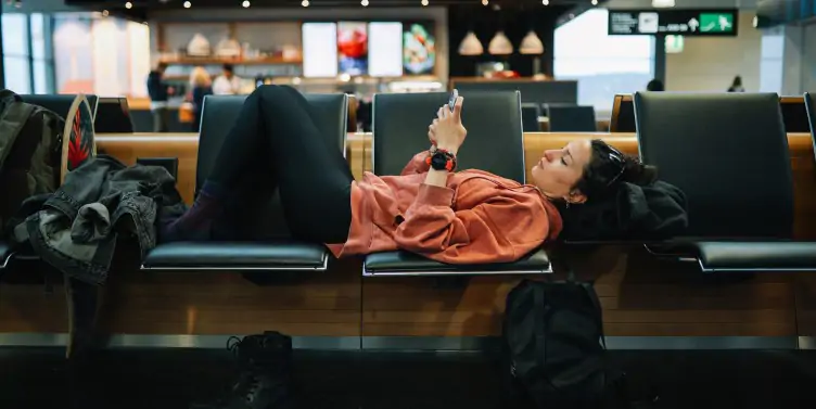 Woman lying across three seats in an airport, reading stuff on her smartphone, waiting for her delayed flight.