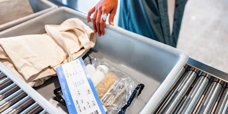 Grey tray filled with 100ml liquid toiletries in a clear bag, boarding pass and a beige tote bag. A female hand is holding the tray on a roller conveyors table at airport security.