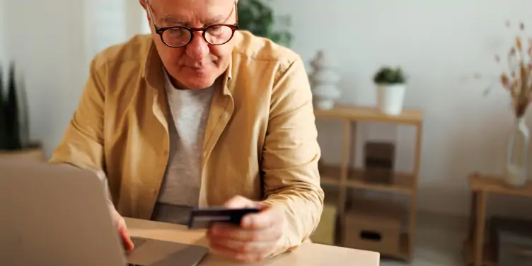 Older gentleman sat at home holding credit card while using laptop for booking a holiday online