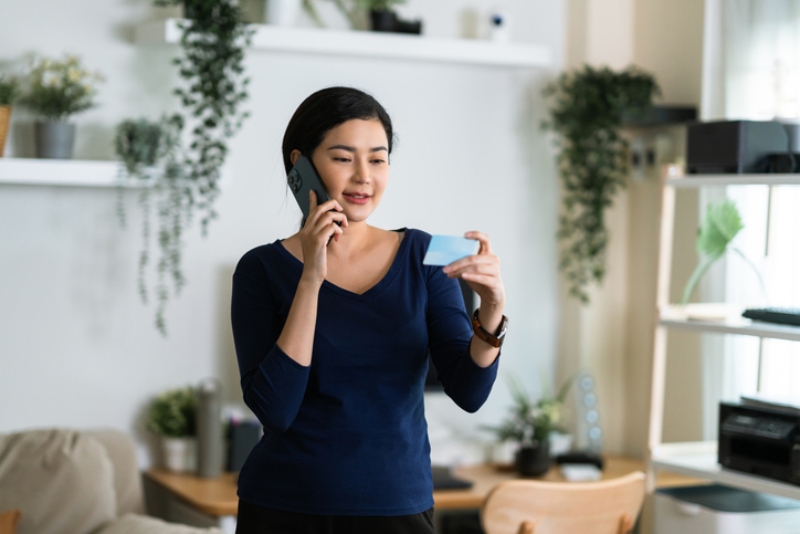 Engaged in a conversation, a woman talks on the phone while holding her insurance ID card, managing her healthcare in a bright, plant-filled room.