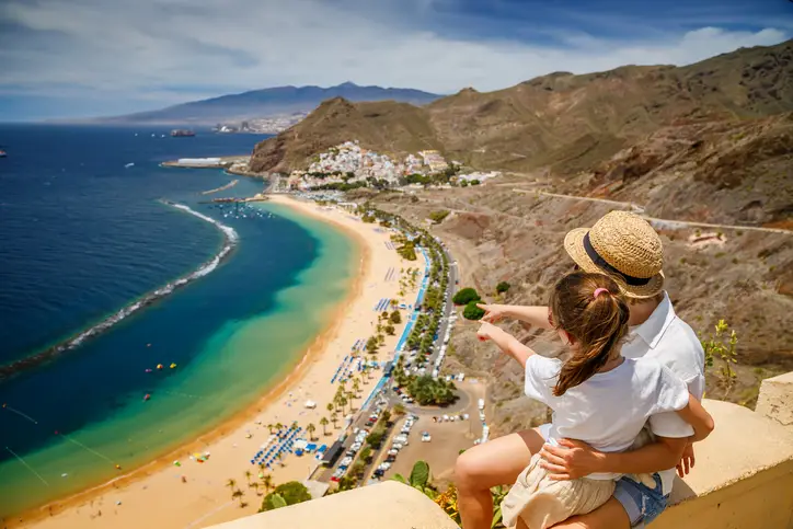 Mother and daughter sitting on a ledge, looking out over Las Teresitas beach in Tenerife, pointing towards the beach. Traveling with children concept.